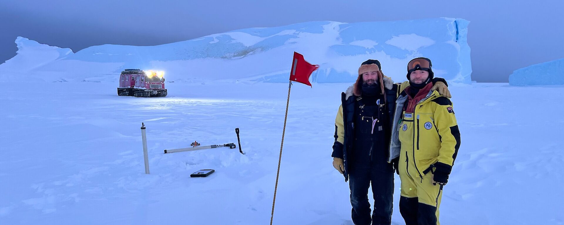 Two people in warm clothing stand proudly next to a red flag and some equipment on the sea ice, with a pink vehicle and a large iceberg visible in the distance