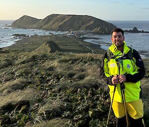 A smiling man wearing bright yellow outdoor gear stands on top of a grassy hill that overlooks an island isthmus and a large hill in the background. The ocean surrounds the landscape.
