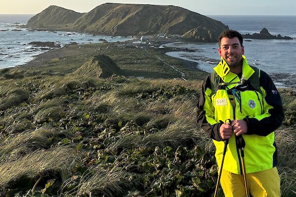 A smiling man wearing bright yellow outdoor gear stands on top of a grassy hill that overlooks an island isthmus and a large hill in the background. The ocean surrounds the landscape.