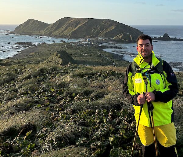 A smiling man wearing bright yellow outdoor gear stands on top of a grassy hill that overlooks an island isthmus and a large hill in the background. The ocean surrounds the landscape.