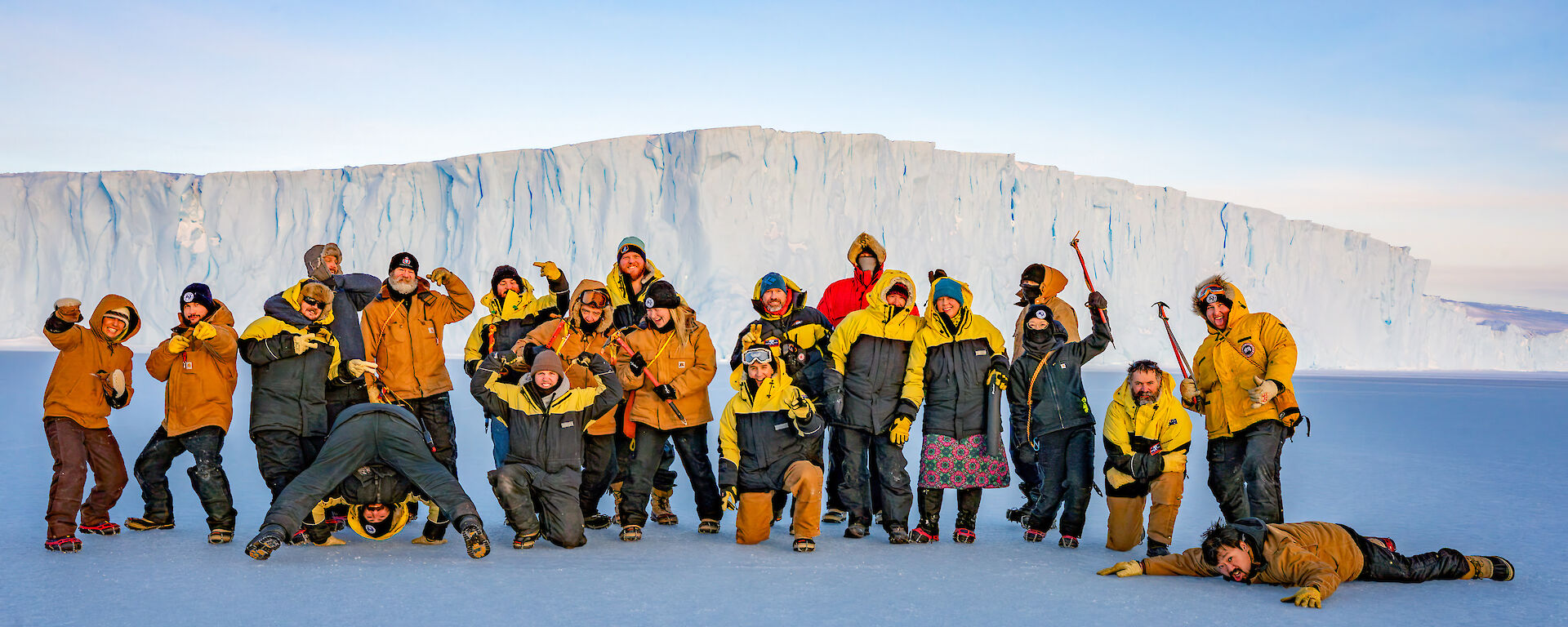 A large group of expeditioners stiking amusing poses in front of an ice cliff.
