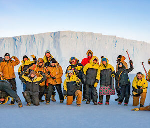A large group of expeditioners stiking amusing poses in front of an ice cliff.