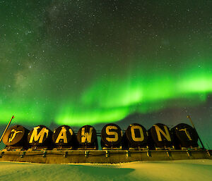 The Mawson Station sign under a vivid green aurora in the night sky.