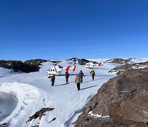 Five people approach a helicopter sitting on a flat patch of ice, surrounded by rocky outcrops.