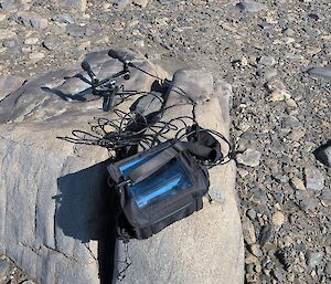 A black box with a number of cables stretching to two microphones sits on a large brown rock.