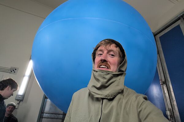 A man wearing a khaki brown protective jacket is standing in front of a large blue balloon