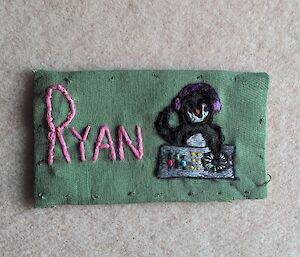 A rectangular green fabric name tag contains the name Ryan embroidered in pink and an embroidered image of a penguin operating DJ decks