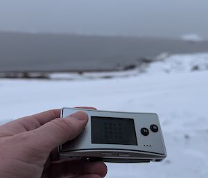 A hand hols a small silver device with a black screen and small black buttons. It is on a snowy beach in front of calm water