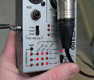 A hand holds a silver box with a large black cable coming out of it and a number of red lights and black switches