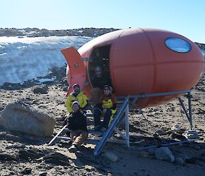 An orange fibreglass hut, shaped like a disk with a ladder, on rocky ground with ice in the background