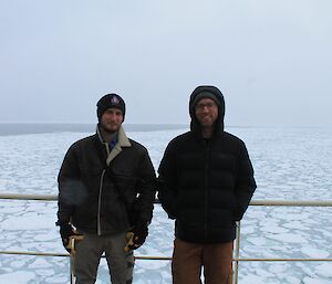 Two men, wearing warm clothing stand side by side in front of a railing, with ice water visible behind.