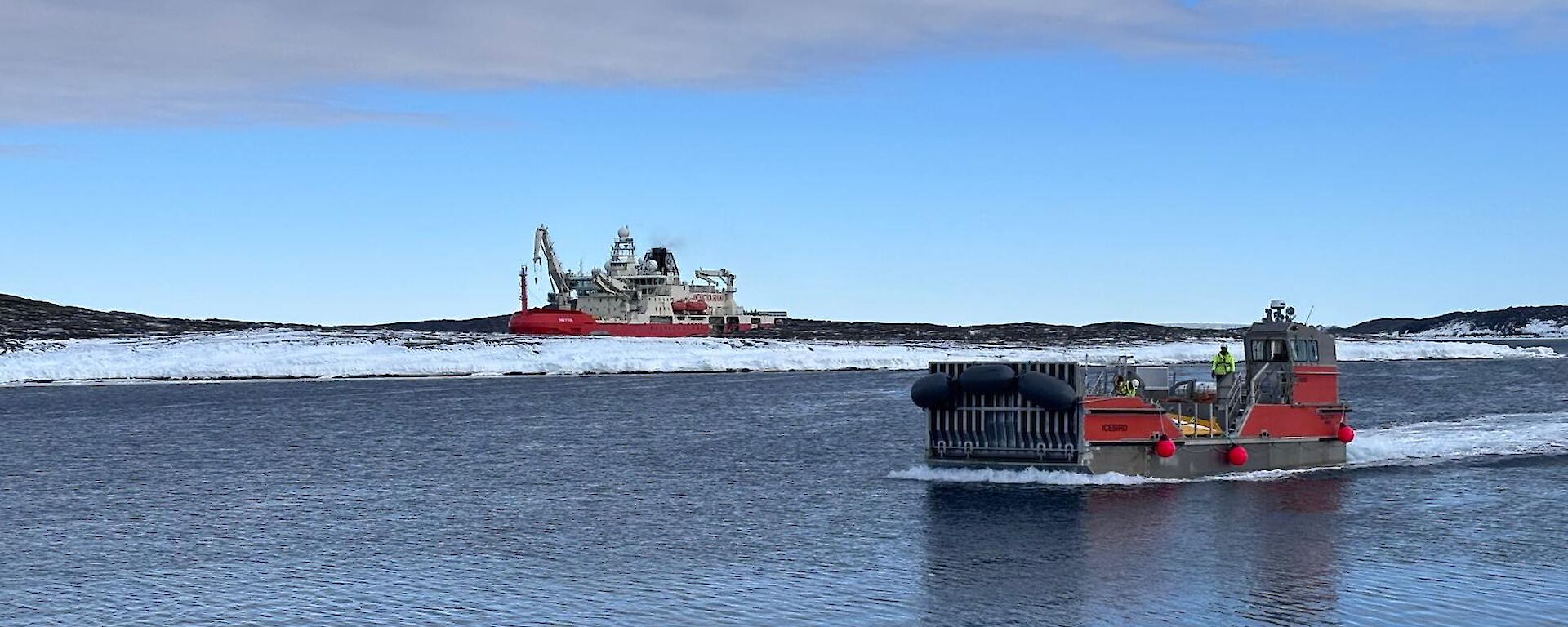 A red barge on water in the foreground with the icebreaker Nuyina in the background