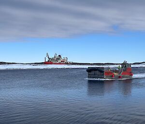 A red barge on water in the foreground with the icebreaker Nuyina in the background