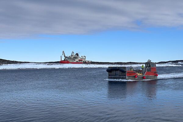 A red barge on water in the foreground with the icebreaker Nuyina in the background