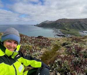 A smiling woman wearing a bright yellow outdoor jacket and blue and grey beanie sits atop a hill while looking down at an island isthmus and hilly terrain surrounded by ocean.