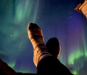 A green aurora dances across a star-lit sky as viewed between the sock-clad feet of someone sitting down with their feet up.