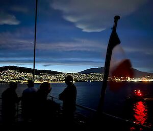 Just after dusk, people standing on a ships deck where a French Flag is being flown. There are city lights in the background on the distant shoreline.
