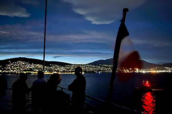 Just after dusk, people standing on a ships deck where a French Flag is being flown. There are city lights in the background on the distant shoreline.