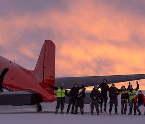 A group of expeditioners standing behind a stationary red aircraft and waving