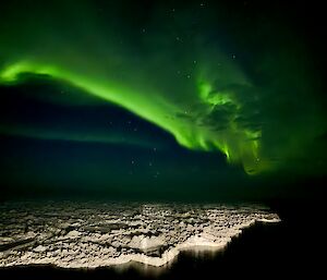 Nuyina lights up the passing sea ice at night with a green aurora in the background
