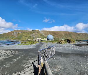 A sandy isthmus with ocean on the left side and a large, mountainous green plateau in the distance with blue sky and clouds above it.