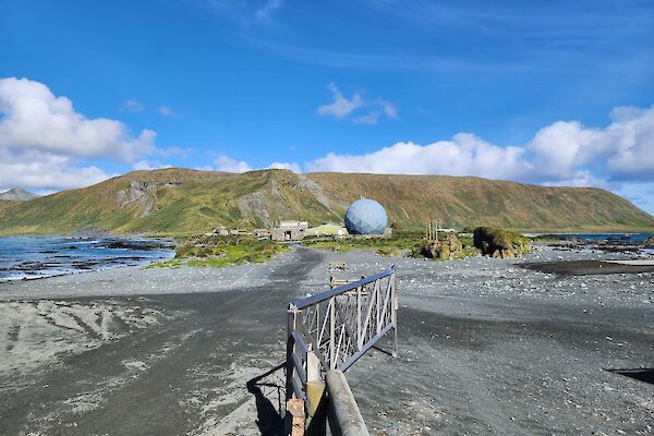 A sandy isthmus with ocean on the left side and a large, mountainous green plateau in the distance with blue sky and clouds above it.