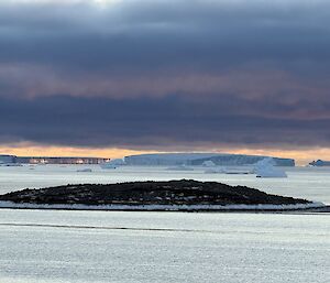 A small rocky island with a number of icebergs in the background. On the left hand side of the picture the wall of one iceberg, C39, is reflecting the orange glow of the sunset.