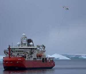 A white helicopter with a long line attached underneath leaves a large white and red ship that is parked in still waters with icebergs in the background.