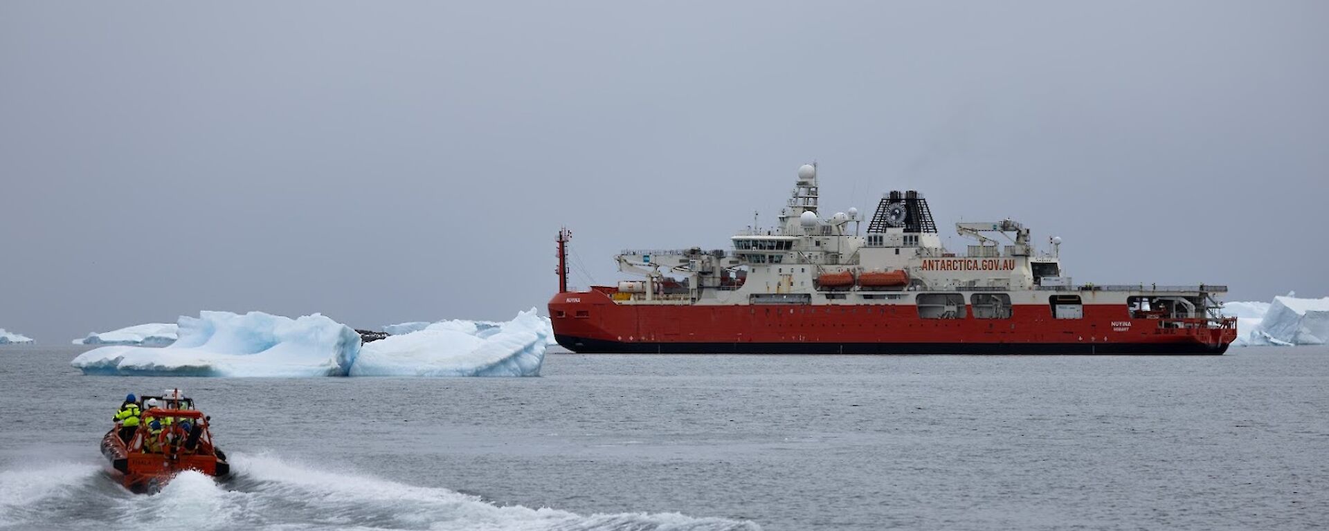 A smaller orange boat moves through the water to a large red and white vessel with people in high visibility clothing on board.