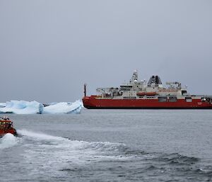 A smaller orange boat moves through the water to a large red and white vessel with people in high visibility clothing on board.