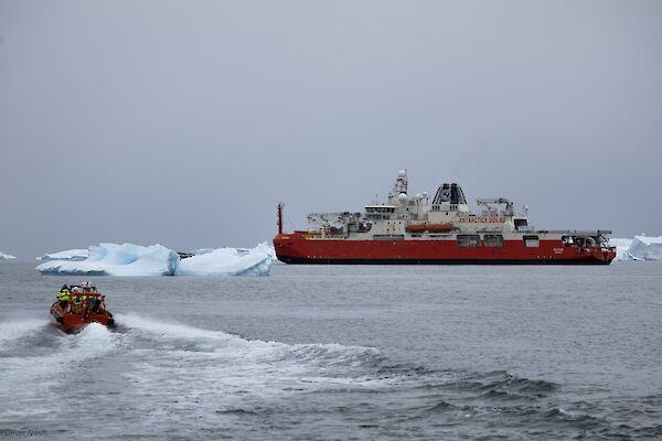 A smaller orange boat moves through the water to a large red and white vessel with people in high visibility clothing on board.