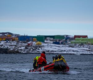 A small inflatable boat with 7 people aboard wearing safety clothing heads towards the camera, in the background are multiple coloured buildings, making up Davis Station