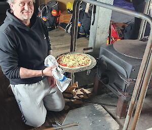 A man holding an uncooked pizza in front of a pizza oven in a shed