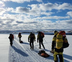 Six people in Antarctic yellow outfits walking along a snowy plain, back to the camera, under a blue sky with white clouds
