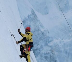 A person in Antarctic yellows climbing a steep ice cliff with two ice axes