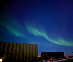 A wispy green aurora angles up into a deep blue sy, with some stars visible. In the foreground are yellow and green buildings.