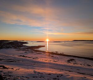 The sun is just about to dip below the horizon, with the sky blue and orange, with come clouds. the golden colour is reflected in the snow on the shore.