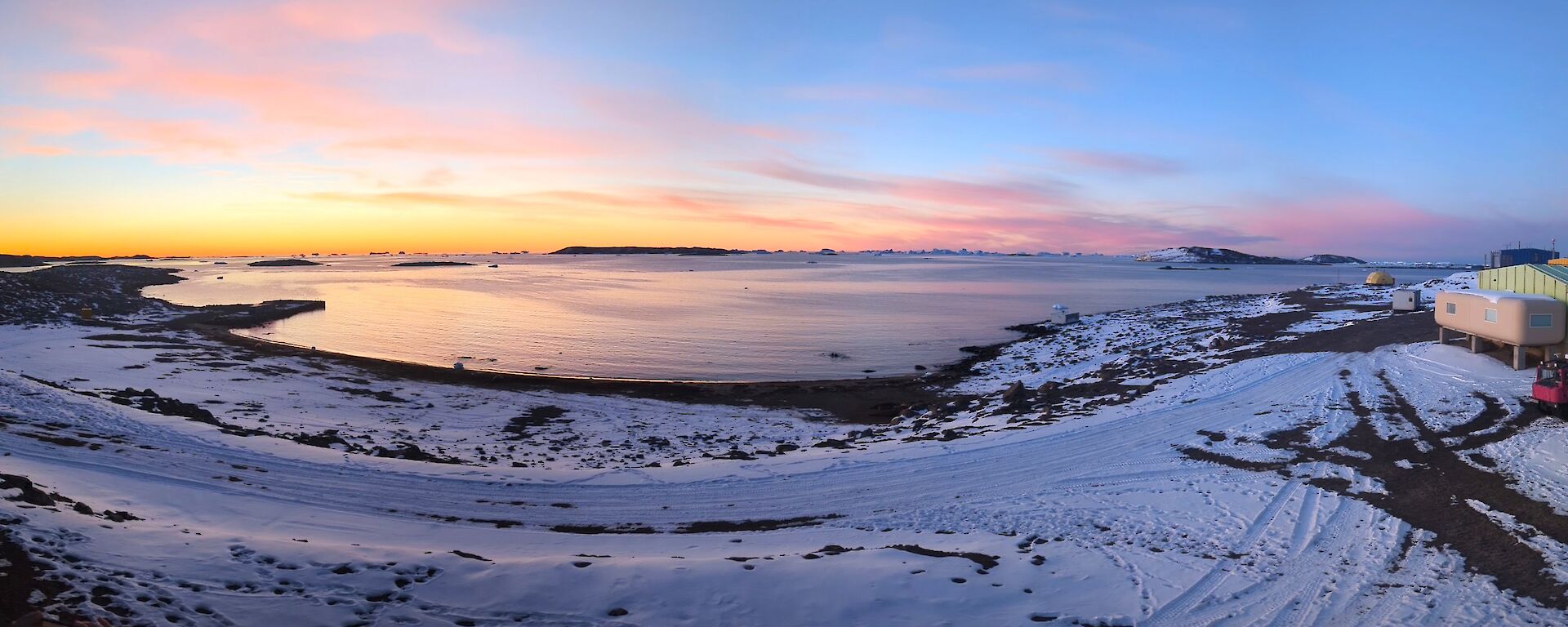 A wider, panoramic shot shows a snowy landscape curling around a bay. The sun is setting to the left of the picture, with a layer or bright orange on the horizon, some pink wispy clouds and a bold blue sky above