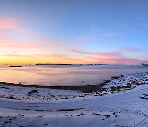 A wider, panoramic shot shows a snowy landscape curling around a bay. The sun is setting to the left of the picture, with a layer or bright orange on the horizon, some pink wispy clouds and a bold blue sky above