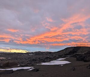 A rocky landscape with a cloudy sky above, reflecting a bold orange sunset.