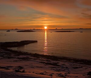 The sun is about to set over water, with dep oranges and browns filling the slightly cloudy sky and landscape
