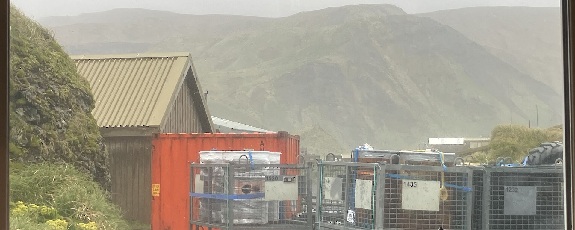 A large window through which can be seen stacked cage pallets, an orange sea container and a rugged green plateau in the distance.