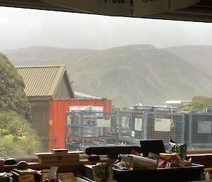 A large window through which can be seen stacked cage pallets, an orange sea container and a rugged green plateau in the distance.