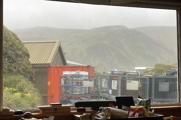 A large window through which can be seen stacked cage pallets, an orange sea container and a rugged green plateau in the distance.
