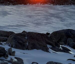 The sun setting behind dark hills with ice and black rocks in the foreground