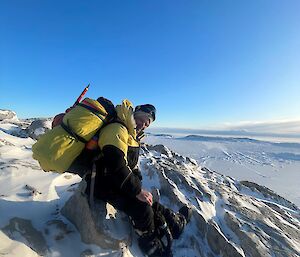 A smiling man wearing heavy snow gear and carrying a large backpack and ice-axe, sits on an icy ridge in a snow covered landscape.