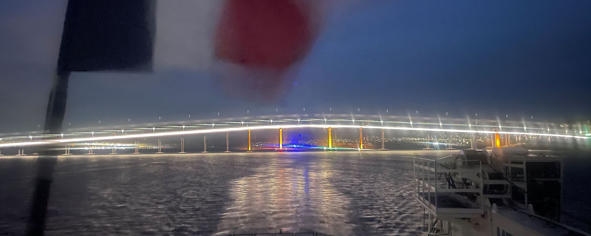 The back helicopter deck of a ship flying a French flag, at nighttime, looking towards a large arched traffic bridge that is well lit up and reflecting off the water.