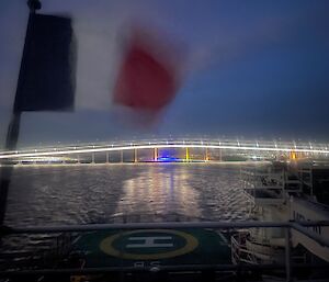 The back helicopter deck of a ship flying a French flag, at nighttime, looking towards a large arched traffic bridge that is well lit up and reflecting off the water.