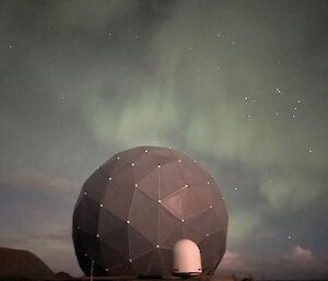 A dark grey golf-ball shaped dome about 10m high set against the night sky with a green aurora and many stars.