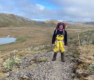 A smiling man wearing hiking gear and carrying a large red backpack stands on a track set in a green, hilly landscape.